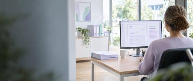Mitarbeiterin im Büro prüft am Bildschirm ein digitales Dokument, neben dem Monitor liegt ein Stapel Papierbelege (AI generated)